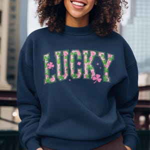 May include: Navy blue sweatshirt with the word "LUCKY" in green and pink lettering. The letters are filled with a shamrock pattern. Pink shamrocks and stars are scattered around the word.