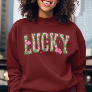 May include: A maroon sweatshirt with the word "LUCKY" in large, stylized letters across the chest. The letters are filled with a pattern of pink shamrocks and green leaves, with pink stars and shamrocks around the letters.