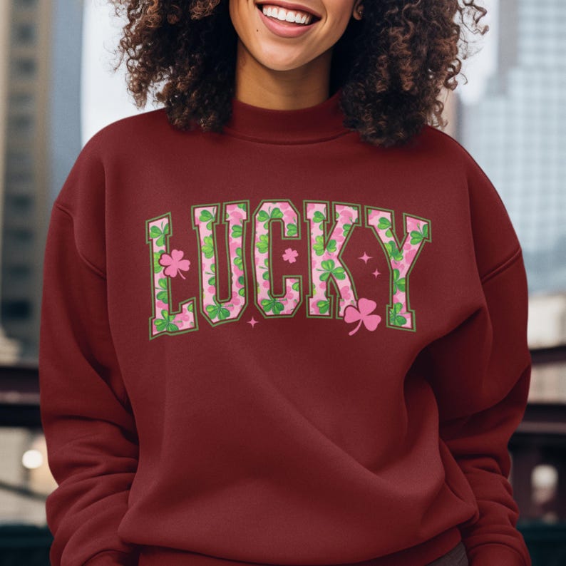 May include: A maroon sweatshirt with the word "LUCKY" in large, stylized letters across the chest. The letters are filled with a pattern of pink shamrocks and green leaves, with pink stars and shamrocks around the letters.