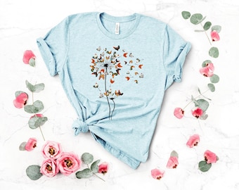 May include: A light blue t-shirt with a dandelion design made of butterflies. The butterflies are in shades of orange, brown, and white.
