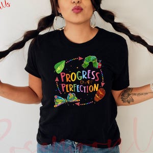 Puede incluir: Camiseta negra con un diseño gráfico colorido que presenta una oruga, una mariposa y el texto "Progress over Perfection".