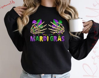 May include: Black sweatshirt featuring a skeleton hand holding a purple and green Mardi Gras fleur-de-lis. The text "Mardi Gras" is written in green, purple, and yellow.