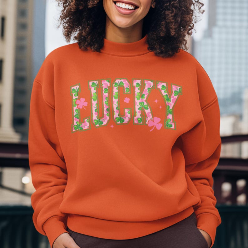 May include: An orange sweatshirt with the word "LUCKY" in a green and pink clover design. The sweatshirt has a crew neck and long sleeves. The image is taken outdoors.