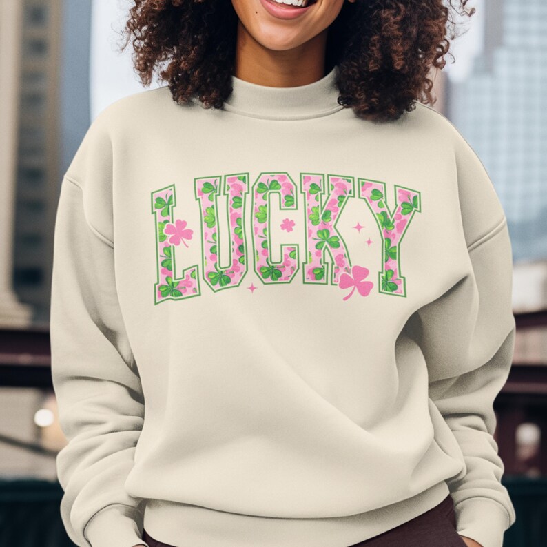 May include: Cream-colored sweatshirt with the word "LUCKY" in green outlined letters filled with pink and green shamrock patterns. The sweatshirt has a crew neck and long sleeves, perfect for St. Patrick's Day.