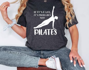 May include: A dark gray t-shirt with a white graphic that says "If it's easy, it's probably not Pilates" with a silhouette of a person doing a Pilates exercise.