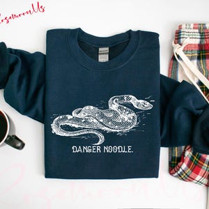 May include: Navy blue sweatshirt featuring a white illustration of a snake with the text "DANGER NOODLE." A red and black coffee mug and plaid pajama pants are also visible. The sweatshirt is folded, with the sleeves draped over the other items.