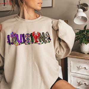 May include: A beige sweatshirt with a purple, green, and gold design that says "LOUISIANA". The design includes a Mardi Gras mask, a crawfish, and beads.