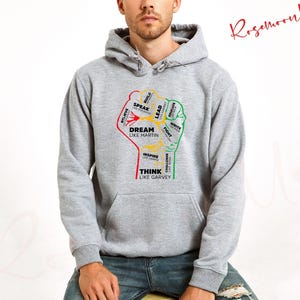 May include: Gray hooded sweatshirt with a raised fist graphic. The fist is filled with words like "Dream Like Martin", "Think Like Garvey", "Speak", "Lead", "Inspire", "Challenge", and more.