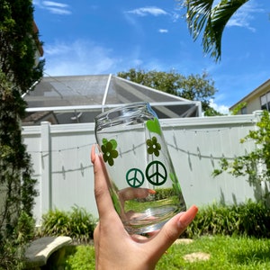 May include: A clear glass with green peace signs and flowers. The glass is being held by a hand.