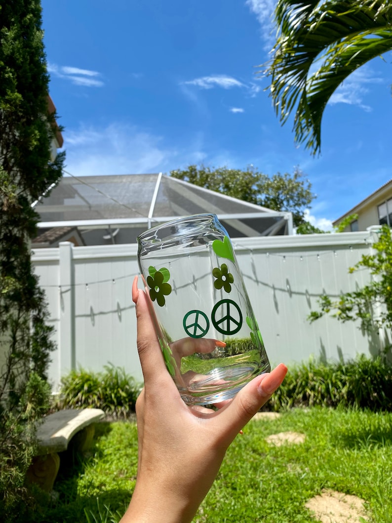 May include: A clear glass with green peace signs and flowers. The glass is being held by a hand.