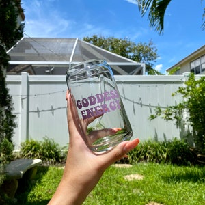 May include: A clear glass cup with a pink "Goddess Energy" graphic on the side. The cup is being held by a hand in a backyard setting.