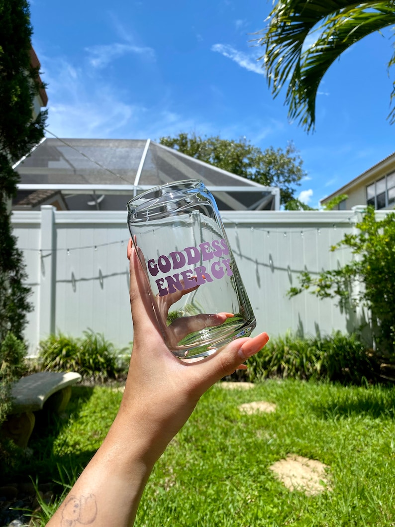 May include: A clear glass cup with a pink "Goddess Energy" graphic on the side. The cup is being held by a hand in a backyard setting.