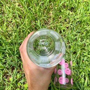 May include: A clear glass water bottle with a pink smiley face design. The bottle has the words "Smile Today" printed on the bottom.