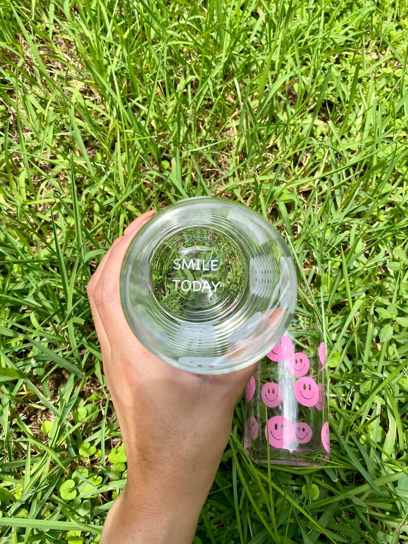 May include: A clear glass water bottle with a pink smiley face design. The bottle has the words "Smile Today" printed on the bottom.