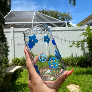 May include: A clear glass can with blue peace signs and flowers. The can is being held by a hand.