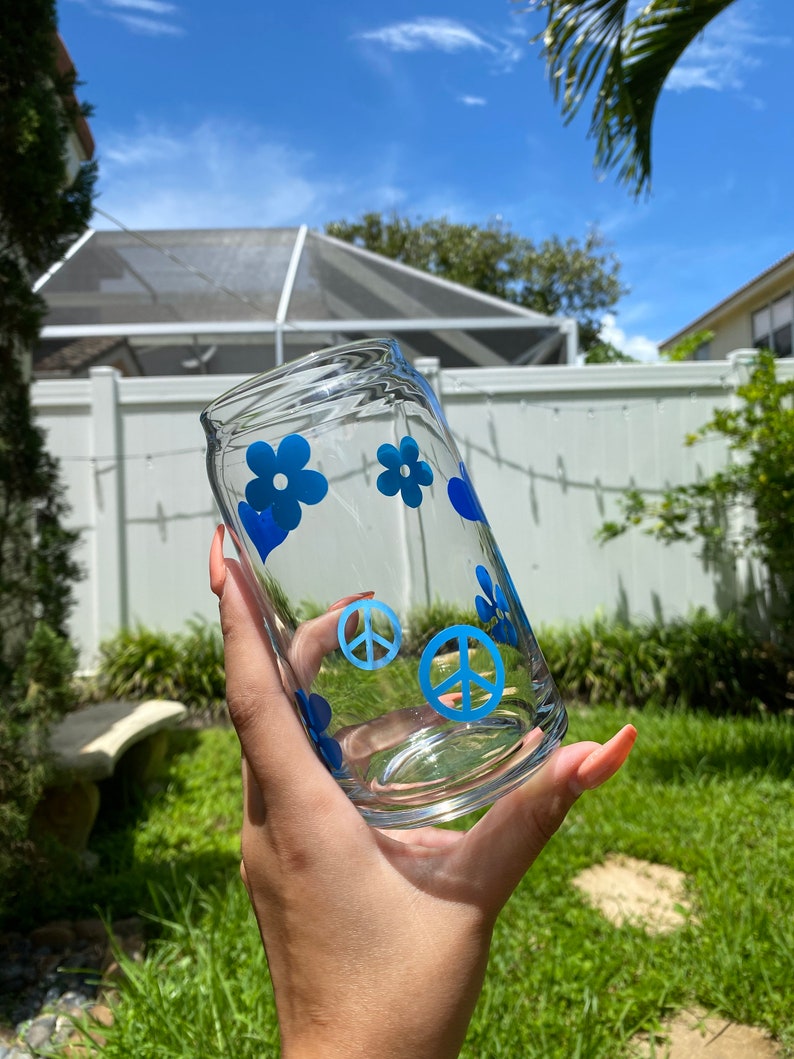 May include: A clear glass can with blue peace signs and flowers. The can is being held by a hand.