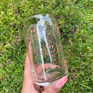 May include: A clear glass tumbler with a pink smiley face pattern. The tumbler is held in a hand with pink nail polish.