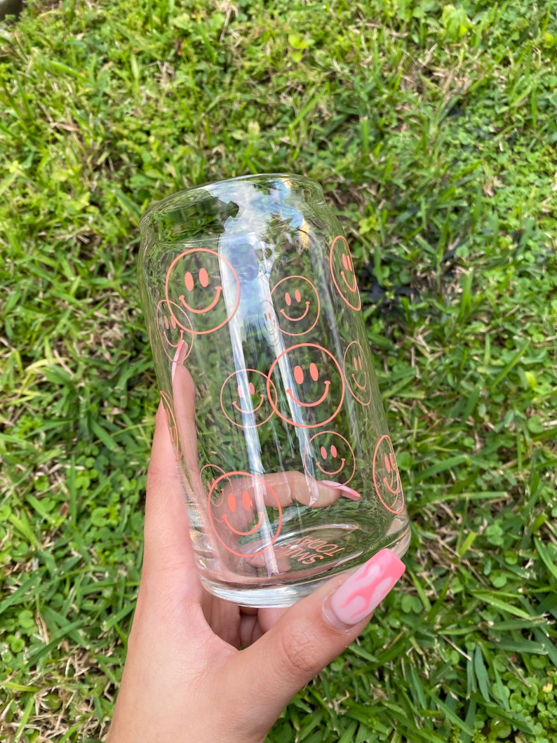 May include: A clear glass tumbler with a pink smiley face pattern. The tumbler is held in a hand with pink nail polish.