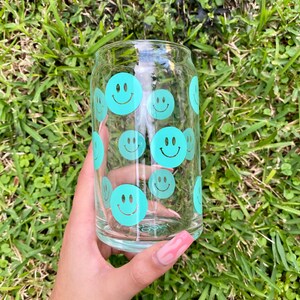 May include: A clear glass can with a teal smiley face pattern. The can is held in a hand with pink nail polish.