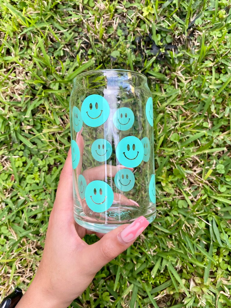 May include: A clear glass can with a teal smiley face pattern. The can is held in a hand with pink nail polish.