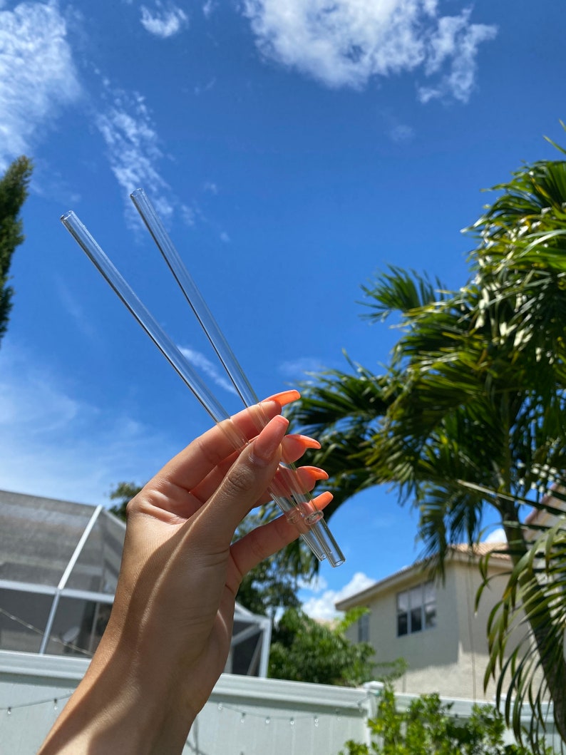 May include: Two clear glass drinking straws held in a hand against a blue sky background.