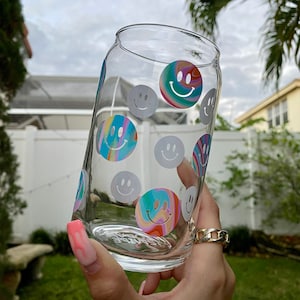 May include: A clear glass can with a rainbow swirl design and white smiley faces. The can is being held by a hand wearing a gold chain ring.