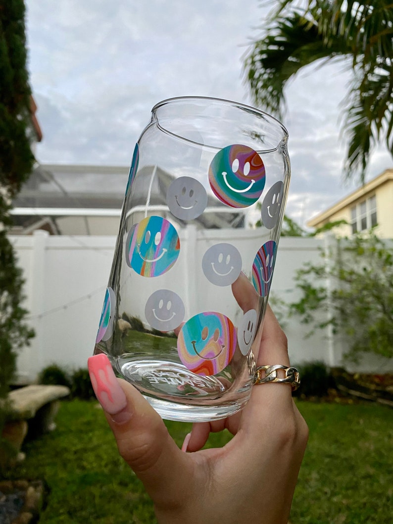 May include: A clear glass can with a rainbow swirl design and white smiley faces. The can is being held by a hand wearing a gold chain ring.