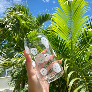 May include: A clear glass can with a white smiley face design. The can is being held by a hand against a background of green palm leaves and a blue sky.