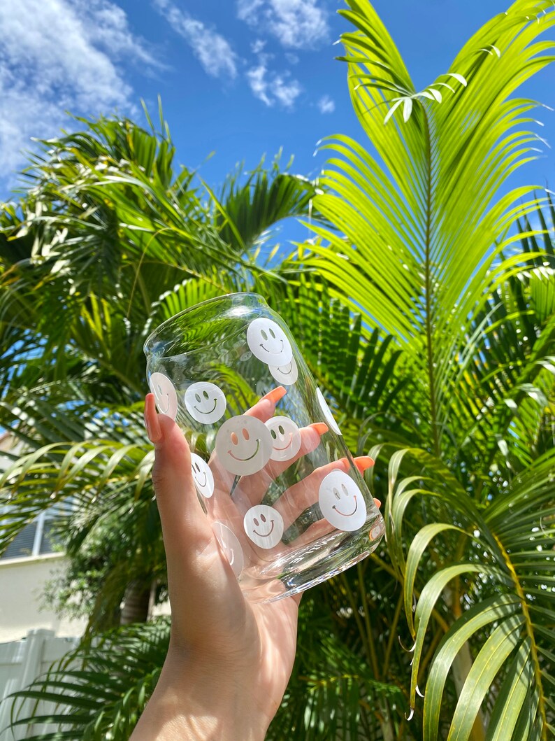 May include: A clear glass can with a white smiley face design. The can is being held by a hand against a background of green palm leaves and a blue sky.