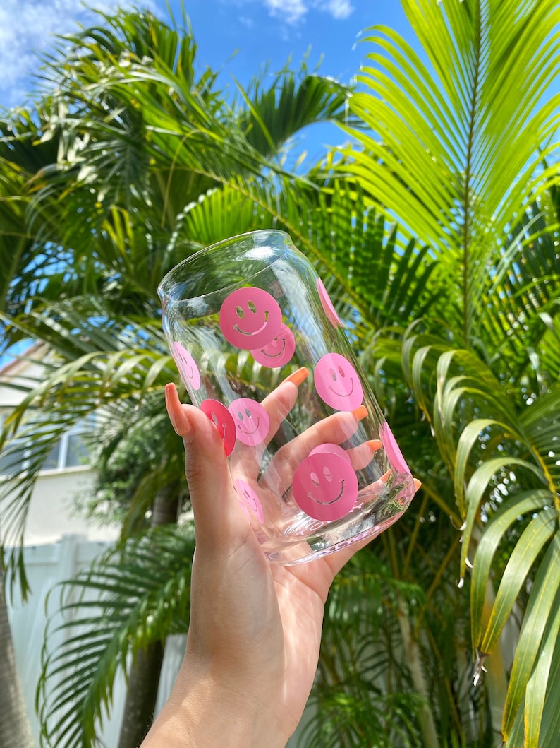 May include: A clear glass with pink smiley faces printed on it. The glass is being held by a hand against a background of green palm trees and a blue sky.