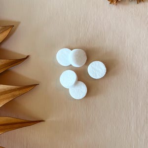 May include: A collection of small, white, circular felt pads arranged on a light brown surface. The pads are clustered together, suggesting a craft or DIY project. Dried floral elements are in the background.