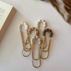May include: A collection of five gold-colored paper clips, each adorned with a macrame knot in varying neutral tones of cream, beige, and brown. The clips are arranged on a white surface, with a book and a tan fabric in the background.