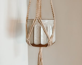 May include: A white ceramic planter with a speckled glaze hangs from a macrame plant hanger. The hanger is made of natural cotton rope and has a tassel at the bottom.