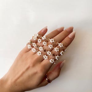 May include: A set of four white beaded bracelets with gold accents, featuring a daisy flower design. The bracelets are stacked on a person's wrist.