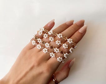 May include: A set of four white beaded bracelets with gold accents, featuring a daisy flower design. The bracelets are stacked on a person's wrist.
