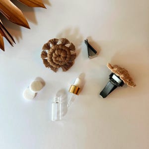 May include: A collection of car air freshener accessories. Includes a brown macrame flower, a black vent clip, a small glass bottle with a gold cap and dropper, two white felt pads, and a macrame air freshener for the car vent.