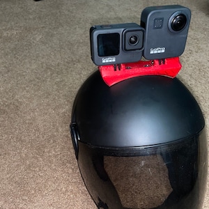 Puede incluir: Dos cámaras GoPro negras montadas en un soporte de casco rojo, fijadas a un casco de moto negro.