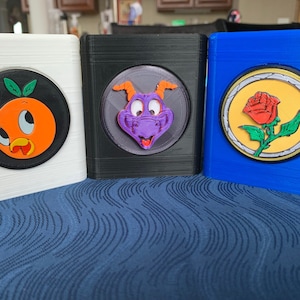 Peut inclure: Trois boîtes imprimées en 3D avec différents motifs de personnages Disney. La première boîte est blanche avec un cercle noir contenant une orange de dessin animé avec un visage d'oiseau. La deuxième boîte est noire avec un cercle gris contenant un dragon violet avec une corne verte. La troisième boîte est bleue avec un cercle jaune contenant une rose rouge avec des feuilles vertes.