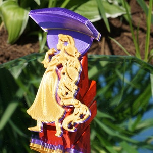 Puede incluir: Una figura impresa en 3D de color dorado de una mujer con un sombrero morado. La figura está de pie sobre una base roja con un borde morado.