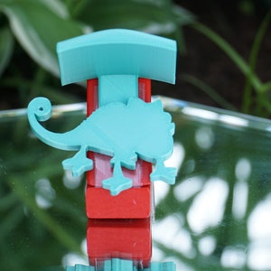 Puede incluir: Una figurita de camaleón azul turquesa sobre una base roja y azul turquesa. La figurita está sobre una superficie de espejo.