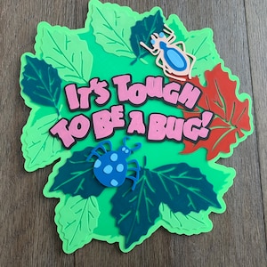 以下が含まれることがあります： 緑、ピンク、青、赤のフォームクラフトで、「It's Tough To Be A Bug!」というテキストが、緑の葉と2匹の虫に囲まれています。