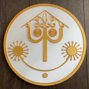 Puede incluir: Un letrero circular blanco con un borde dorado y un diseño dorado en el centro. El diseño presenta una casa estilizada con dos soles a cada lado.