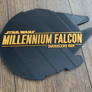 May include: A black Millennium Falcon sign with gold lettering. The sign reads "STAR WARS MILLENNIUM FALCON SMUGGLERS RUN". The sign is shaped like the Millennium Falcon spaceship.