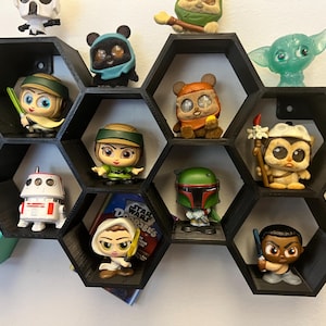 Pode incluir: Uma estante hexagonal preta com 10 compartimentos, exibindo uma variedade de pequenas figuras, incluindo personagens de Star Wars, um robô e uma criatura da Lagoa Negra. A estante está montada em uma parede branca.