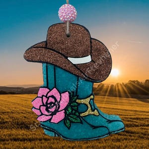 May include: A turquoise cowboy boot and brown cowboy hat ornament with glitter accents. A pink glitter rose and gold buckle detail the boot. A pink glitter ball hangs from a string at the top. The background shows a sunset over a field.