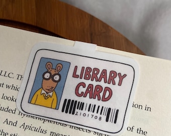 Puede incluir: Una tarjeta de biblioteca blanca con una ilustración de dibujos animados de una persona con gafas y una camisa amarilla. La tarjeta tiene el texto "LIBRARY CARD" en letras negras y un código de barras con el número "2101708".