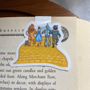 Peut inclure: Un marque-page en forme de route de briques jaunes représentant les personnages du Magicien d'Oz : Dorothy, l'Épouvantail, l'Homme de Fer-Blanc et le Lion peureux. Les personnages marchent sur la route de briques jaunes, qui est représentée dans un style cartoon.
