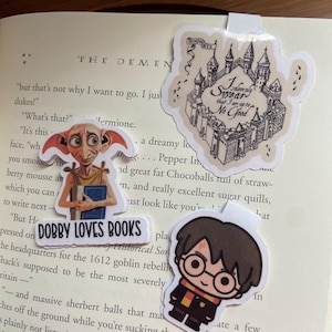 May include: A set of four Harry Potter themed stickers. One sticker features a cartoon image of Harry Potter with his signature glasses and a Gryffindor scarf. Another sticker features a cartoon image of Dobby the house-elf holding a book. The third sticker features a cartoon image of Hogwarts castle with the text "I solemnly swear that I am up to no good." The fourth sticker features a cartoon image of a house-elf with large ears and a book in his hand. The text "Dobby Loves Books" is printed on the sticker.