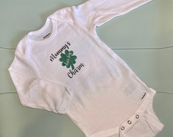 May include: White baby bodysuit with long sleeves. The bodysuit features the text "Mommy's Lucky Charm" in black script with a green shamrock graphic. The garment has snap closures at the bottom and a round neckline.