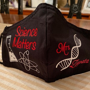 May include: Black fabric face mask with the text "Science Matters" in red lettering and a beaker, atom, and DNA strand in white. The text "Mrs. Zapata" is also in red lettering.